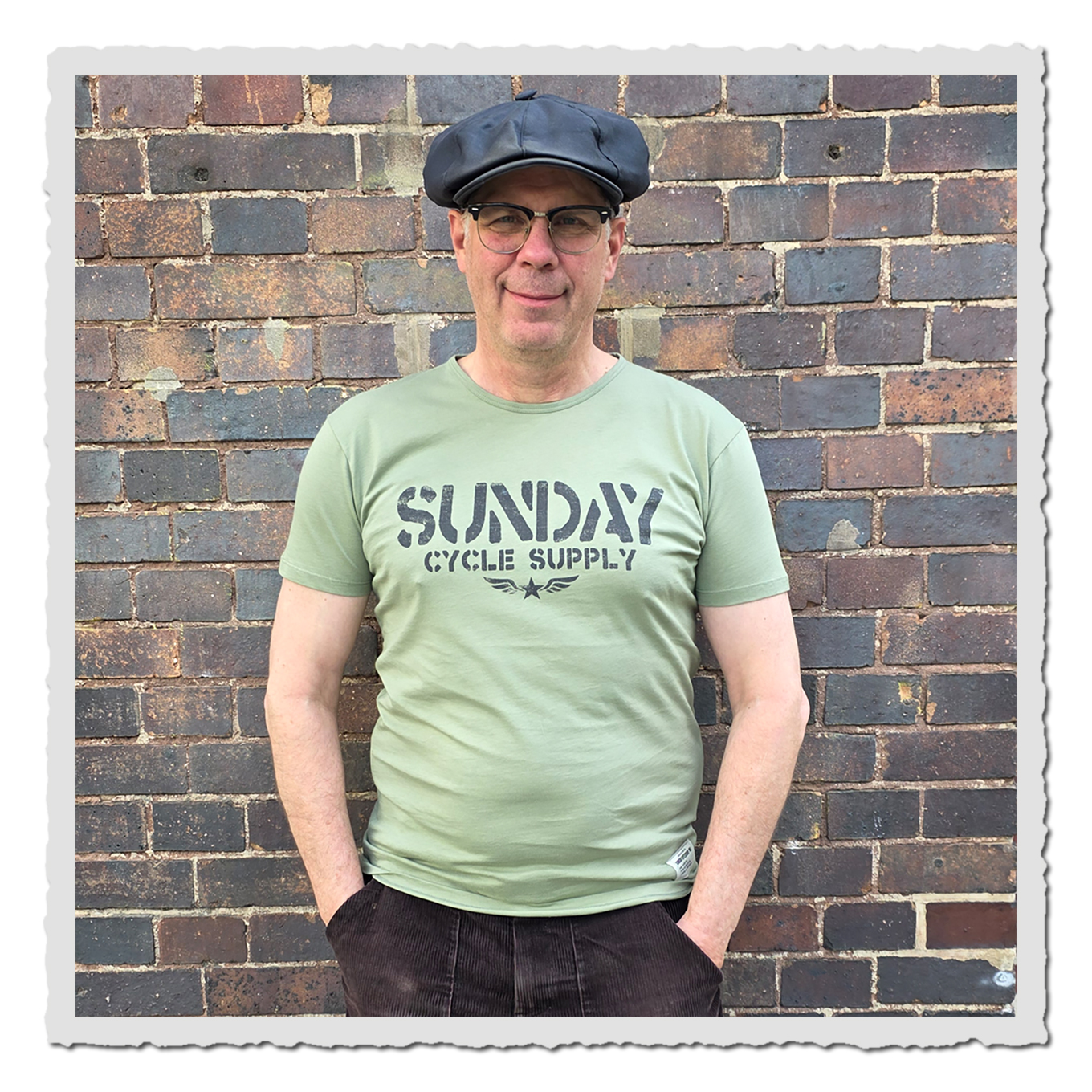 Mann trägt hellgrünes T-Shirt mit dunklem Frontdruck vor einer Backsteinwand, Vorderansicht.